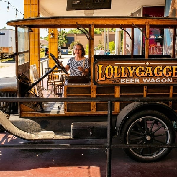 A smiling woman sits in a vintage "Lollygaggers Beer Wagon" vehicle, parked outdoors.