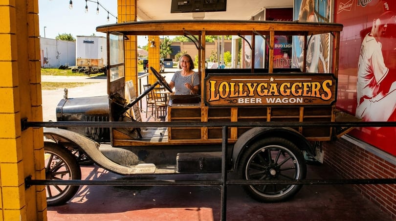 A smiling woman sits in a vintage "Lollygaggers Beer Wagon" vehicle, parked outdoors.