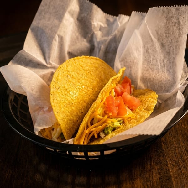 Two hard-shell tacos with cheese, guacamole, and diced tomatoes in a basket with paper.