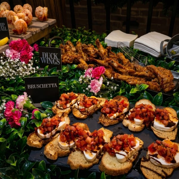 A buffet spread with bruschetta, duck wings, and shrimp cocktail, adorned with pink flowers and greenery.